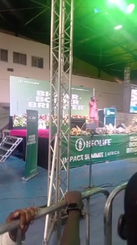 The video shows an indoor event, presumably a NeoLife Impact Summit Africa, with a speaker on stage wearing a pink outfit. Behind the speaker are banners that read "Bigger, Bolder, Brighter" and display the NeoLife logo. A large crowd is present, some of whom are recording the event on their phones. The lighting is bright, with spotlights shining on the stage. The event is taking place in a large hall or stadium-like setting. The camera is positioned behind a metal railing.