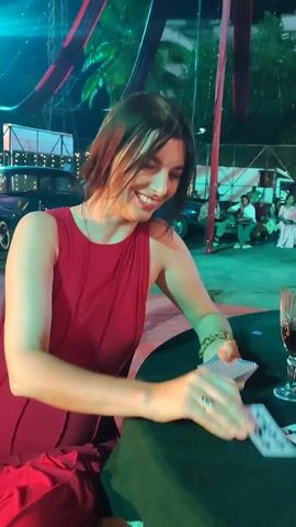 Here is a description of the video:

The video shows a woman in a red dress playing cards at a table. She is sitting at a round table covered with a green cloth. There are three cards face up on the table, all showing the ten of clubs. She is holding a deck of cards in her hands, smiling as she sorts through them.  A glass of dark red liquid is also on the table.  In the background, there are people and a vintage car, with some decorations hanging above. The scene appears to be outdoors at night, with some artificial lighting.