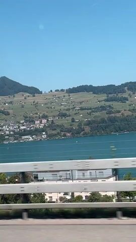 This video shows a view from a vehicle, possibly a train or car, traveling along a road next to a lake. The lake is blue and calm, with a town or village situated along its shores. Behind the town, rolling green hills rise up, dotted with scattered houses and trees. The sky is clear and blue. A white metal railing runs along the bottom of the frame, obscuring the immediate foreground. The camera is moving, causing the landscape to blur slightly, especially in the foreground details.