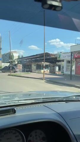 The video appears to be shot from inside a vehicle looking out through the windshield. The viewpoint is low, showing the dashboard in the foreground. Outside, there is a street scene. In the middle ground, there is a building with a damaged or partially destroyed roof and structure. There are some people visible walking around the building. One person, a male wearing dark clothing, walks towards the camera in front of the building. There are cars parked and moving on the street. The environment is urban, with buildings and power lines visible in the background. The weather appears to be sunny with blue skies and some clouds. There is a sign on one of the structures that reads "SKYLINE".