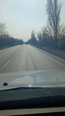 The video shows a first-person perspective from inside a car driving on a straight, paved road. Several other vehicles are visible ahead on the road, driving in the same direction. The sky is pale and overcast. Trees without leaves line both sides of the road. The dashboard of the car is visible at the bottom of the frame. There are white lane markers.