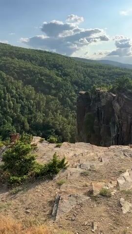 This is a video taken from a high vantage point overlooking a rugged, mountainous landscape. The scene is set during the daytime, with the sun visible in the sky, casting light and shadows across the terrain. The weather appears to be clear with scattered clouds. The foreground is characterized by rocky, uneven ground with sparse vegetation.

The main subject of the video is the dramatic geological formation of a steep cliff face. This cliff is a prominent feature, with layers of rock visible, suggesting a long geological history. Beyond the cliff, the landscape unfolds into rolling hills covered in dense, green forests. In the distance, more mountains are visible, fading into a hazy horizon.

The video captures a panoramic view, panning across the expansive scenery. The overall impression is one of natural beauty and rugged wilderness. The sunlight suggests it's likely late afternoon or early evening, given the angle and warmth of the light. The location context provided is Hrabrino, Bulgaria, which places this scenic view within a specific geographical region. There are no people or man-made objects visible in the video, emphasizing the wild and untamed nature of the setting. The emotions evoked are likely awe and tranquility, inspired by the grandeur of the natural landscape. No text is visible in the video.
