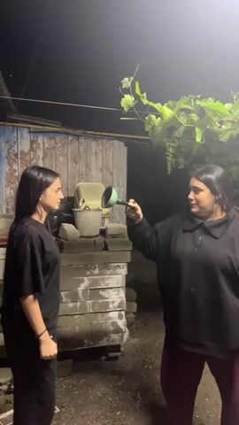 The video begins with two young women standing in what appears to be a backyard at night. The woman on the left, wearing a black t-shirt and black pants, is facing the woman on the right. The woman on the right, wearing a black sweatshirt and burgundy pants, is holding a green watering can and reaching towards the woman on the left. Behind them, there is a wooden structure, a pile of bricks, and a grape vine hanging overhead.

The woman on the right appears to be splashing water from the watering can onto the woman on the left, who then stumbles and falls against a corrugated metal fence. The woman on the left then slides down the fence and lands on the ground. The woman on the right watches her fall and then turns to face the camera with a smile. The scene is dimly lit by artificial lights.