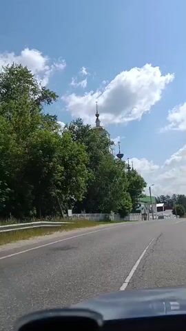 The video shows a view from inside a vehicle, looking forward and slightly to the left. The vehicle is on a paved road with a white dividing line. To the left of the road, there is a white fence and then a white building with green roofs and multiple domes, appearing to be a church. Tall green trees are visible behind the fence and around the church. The sky is blue with scattered white clouds. The video captures the vehicle moving forward, passing the church.