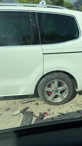 The video shows a white car with significant front-end damage. The front bumper is detached, and the driver's side front panel is crumpled.  Debris is scattered on the ground next to the vehicle. A man in a blue shirt and dark pants walks away from the car, talking on a cell phone. The surrounding environment includes a road with greenery on both sides and a view of distant mountains. The sky is overcast.