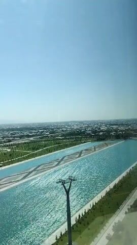 A drone shot pans over a wide, bright blue canal filled with shimmering water. The canal is flanked by manicured green lawns and a paved walkway. In the distance, a city sprawls under a clear blue sky. The scene appears to be a park or recreational area.