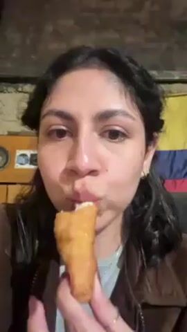 A person with long dark hair held back by a braid is eating a fried snack. The snack appears to be long with a golden brown crispy exterior and filled with molten white cheese which can be seen stretching as the person bites into it. The person is wearing a brown jacket over a gray shirt and a silver necklace. A Colombian flag is visible in the background along with an old-fashioned radio and a wall of rough hewn planks. The room seems dimly lit. The video focuses on the person's face as they eat the snack and smile slightly.