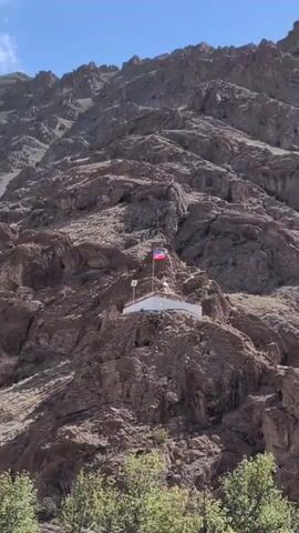 A medium shot shows a shrine built on a steep, rocky mountainside. A national flag flies above the shrine, which is white with some colorful decorations. The mountain is barren and covered in rocks. In the foreground, there are some green trees. The sky is blue with a few wispy clouds.