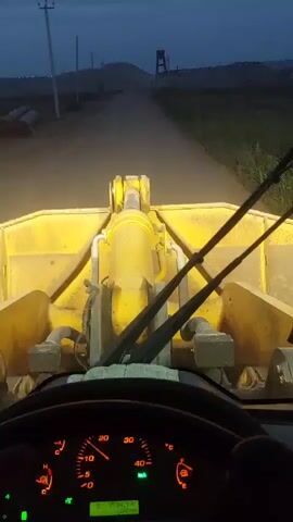 The video begins with a view from inside the cab of a yellow front-end loader. The dashboard instruments are illuminated, showing a speedometer with the needle around 10. The loader's headlights are on, illuminating a dirt road ahead in what appears to be twilight. The loader's bucket and hydraulic arms are visible. The video then pans to the right, showing a side mirror reflecting the interior of the cab, and then a glimpse of the exterior, including some green foliage. The scene transitions to a view of a body of water, possibly a lake, with hills in the background and a bright light reflecting on the water. The sky is overcast. The video appears to be shot from a moving vehicle.