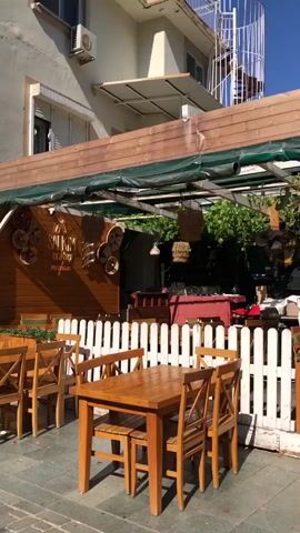 The video shows an outdoor seating area of a restaurant. In the foreground, there are wooden tables and chairs. The tables are set with chairs around them, and some chairs are tucked under the tables. A white picket fence separates the seating area from the rest of the establishment. In the background, there are more tables and chairs, as well as some greenery and decorative elements hanging from the ceiling. The overall impression is a pleasant, relaxed outdoor dining space.