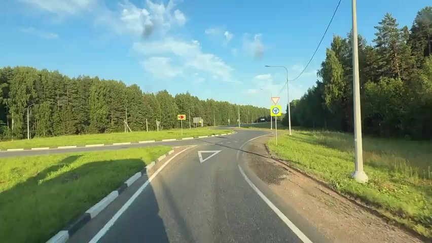 This is a first-person perspective video captured while driving on a paved road. The road appears to be a two-lane road with a white line separating the lanes and a white dashed line at an intersection. On the left side of the road, there is a grassy area with a white curb. On the right side, there is also a grassy shoulder with some patches of dirt. The road is surrounded by a dense forest of green trees. The sky is blue with some scattered white clouds, and the sun is shining, casting shadows on the road. There are several road signs visible. On the left, there is a yellow sign with a black arrow pointing to the left and up, indicating a sharp turn ahead. Further ahead on the left, there is a white square sign with a yellow border and a black arrow. On the right side of the road, there is a pole with two signs. The upper sign is a red and white triangular sign pointing downwards, indicating yield or give way. Below it is a yellow square sign with a blue circular arrow, indicating a roundabout. The vehicle is moving forward and approaching the intersection.