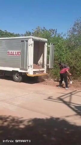 The video shows a white truck with the word "SALUD" written on the side. The back doors of the truck are open and three men are using shovels to unload the truck. The truck is parked on a dirt road with green vegetation in the background. The sky is clear and blue. One man wears a red shirt, another wears a gray shirt and the third wears a blue shirt with colorful shorts. The video is taken during the day.