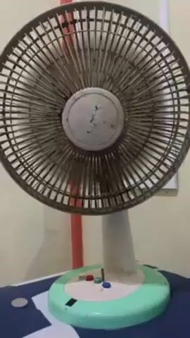 A short video shows a table fan in front of a pale yellow wall. The fan is old and dusty, with a grey grill. The base is white and green and has three buttons on it. The camera pans down to show a table with a blue and white patterned surface. On the table are various objects: a phone charger plugged into an outlet, a small green electric drill, a black hair trimmer with attachments, and a coin. The objects appear to be scattered on the table.