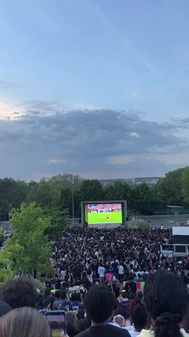 A crowd of people is gathered outdoors to watch a soccer game on a large screen. The screen is positioned on a raised platform, with green trees and the sky in the background. The crowd is dense, with people of various ages and ethnicities visible. Many people are looking towards the screen, while some are holding up their phones to record the event. The sky is partly cloudy with a hint of blue, suggesting it is late afternoon or early evening. A contrail from an airplane is visible in the sky. As the soccer player on screen makes a move, the crowd starts to cheer excitedly.
