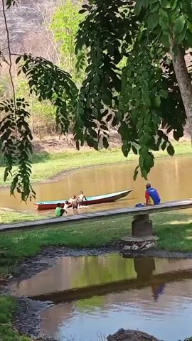 A group of people are in and around a small boat on a muddy river. Some people are in the boat, some are in the water, and one person is sitting on a wooden pier. The water is calm and reflects the surrounding trees and sky. The scene appears to be in a natural, outdoor environment with trees and hills in the background.