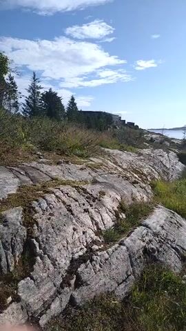 The video shows a view of a rural landscape on a sunny day. In the foreground, there are large, grey rocks with patches of moss and green grass growing between them. A dirt road curves through the scene, leading towards a body of water in the distance. Along the road, there are several houses and some farm-like structures, including what appears to be a yurt. The sky is blue with a few white clouds. Trees are visible on a hill in the background. The sun is shining brightly, creating a lens flare effect in the video. The overall impression is one of a serene and isolated coastal environment.