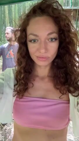 A woman with curly auburn hair and light green eyes, wearing a pink strapless top and a white shirt, is holding a camera at arm's length and recording a selfie video. Behind her are three men; one is standing and holding a camera, another one is standing, and the third is leaning back against a bamboo stalk. They are standing in a bamboo forest. The ground is covered in dry leaves.