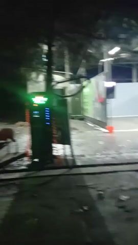 The video shows a self-service car wash at night. The area is lit by overhead fluorescent lights. There are several stalls, separated by grey walls, each equipped with car washing equipment. The ground is wet. The video appears to be taken from outside the car wash area, focusing on the equipment and the stalls. There is a payment kiosk on the left side of the screen. A tree is visible on the right side of the scene.