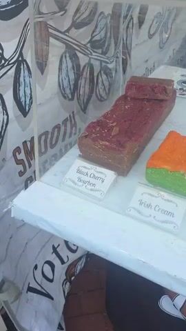 The video shows a display case filled with various types of fudge. The fudge pieces are large, rectangular blocks with different colors and textures, suggesting a variety of flavors. Some pieces are labeled with names like "Black Cherry Bourbon," "Irish Cream," "Penuche," "Salted Caramel Bourbon," and "Creme Brulee." The display is set up outdoors, possibly at a market or fair, as indicated by the brick-like ground visible in the lower portion of the video and a banner that appears to say "MOO." A person's hand is briefly visible in the background, reaching towards something behind the fudge display. The lighting suggests it is daytime.