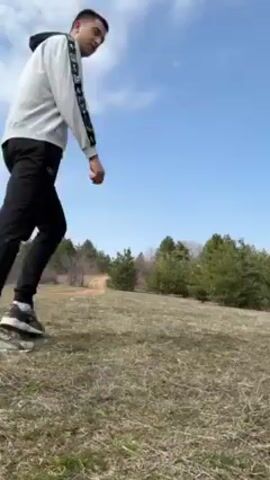 Two people, a man and a woman, are running towards each other on a grassy field under a blue sky with scattered clouds. The man is wearing a white sweatshirt, black pants, and black sneakers. The woman is wearing black leggings, a white long-sleeved shirt, a black vest, and white sneakers. They run towards each other, hold hands, and the man lifts the woman into his arms. In the background, there is a line of trees and a dirt path.