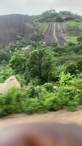 The video shows a rocky mountain covered in greenery, with trees and bushes growing on its slopes. The mountain has a smooth, rounded shape, with some vertical streaks that appear to be worn by water. The sky is cloudy, and the lighting suggests it may be overcast or near sunset. The foreground of the video is blurred, indicating movement, likely from a vehicle. There are some power lines visible on the mountain. The overall impression is of a natural, somewhat wild landscape.