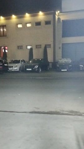 At night, the camera pans across a parking lot in front of a building. The building is a light color, with multiple windows. There are several cars parked in the lot, mostly dark colored sedans and SUVs. In the distance, there are people standing near the street. The ground is light colored concrete. The lighting is provided by the building's exterior lights.