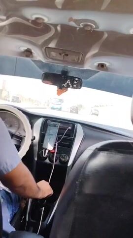 The video shows a person driving a car on a sunny day. The driver is wearing a short-sleeved, button-down shirt. The car's interior is visible, including the steering wheel, dashboard, and rearview mirror. A smartphone is mounted on the dashboard displaying what appears to be a navigation app. Outside the car, other vehicles can be seen on the road. The overall impression is of a routine car ride.