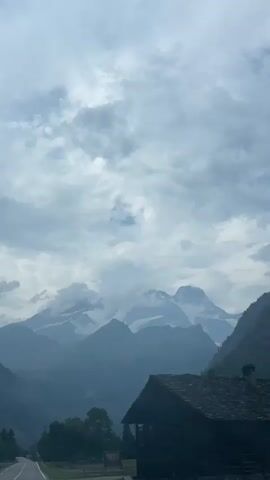 The video shows a perspective from inside a vehicle traveling on a road through a mountainous valley. The sky is overcast with gray clouds. In the foreground, on the right side of the screen, is the dark silhouette of a wooden building with a gabled roof. A road stretches into the distance, curving slightly to the left. On the left side of the road, there are trees and a hint of another vehicle. The middle ground is filled with a dense line of dark green trees. The background is dominated by layers of mountains, with the peaks of the furthest mountains appearing lighter and partially obscured by clouds. The overall atmosphere is somber and serene.
