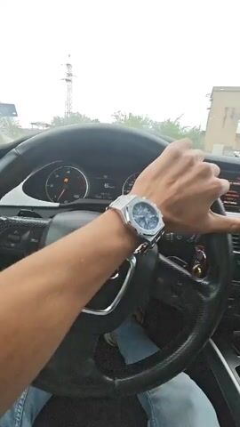 The video shows a first-person perspective of a person driving an Audi car. The driver's hands are visible on the steering wheel, and they are wearing a silver watch on their left wrist. The dashboard and the car's interior are also visible, with the Audi logo prominent on the steering wheel. The view outside the car through the windshield suggests it is daytime and possibly overcast. The driver's legs and feet, clad in blue jeans, are also visible. The video captures the act of driving, with subtle movements of the hands on the steering wheel.