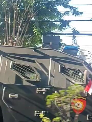 A street scene with several people. The foreground shows the backs of a man carrying a child on his shoulders and a woman in a blue headscarf and dress. In the background, people are gathered on a sidewalk, some holding umbrellas. A large vehicle, possibly a military vehicle, is visible. Trees and power lines are overhead. The Indonesian flag is flying from the vehicle. The overall impression is of a public gathering or event.