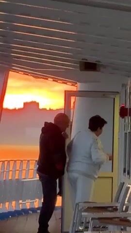The video shows two people on the deck of a vessel. A man wearing a black hoodie and jeans walks away from the camera towards an open doorway. A second person, wearing a white outfit, stands near the doorway, partially obscured by it. The background features a vibrant orange sunset over a body of water, with a city skyline visible in the distance. The deck itself is made of wood, with white railings and a white structure overhead.