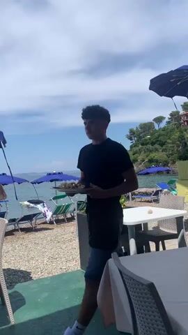 A young man, appearing to be a waiter, walks across a patio area near a beach. He is wearing a black t-shirt, a black apron, and denim shorts. He is holding a tray with food. In the background, there are tables and chairs, blue umbrellas, and the ocean. People are visible on the beach and at tables. The man approaches a table where a man in a white shirt is seated. The waiter is in the process of serving food or placing it on the table. The sky is blue with some clouds.