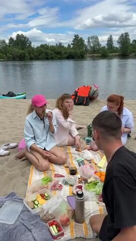 This media captures a sunny day at a sandy beach next to a calm river, likely during the summer in Smolenshchina, Russia. The sky is a vibrant blue with scattered white clouds, suggesting pleasant weather. Several people are enjoying a picnic and spending time together by the water.

In the foreground, a group of four individuals are seated on a blanket, sharing food from an assortment of containers and bags. One person, wearing a light blue shirt and pink hat, is in the process of eating. Another person in a white shirt and pink shorts is also seen with food. A man in a black t-shirt is actively engaged with the food laid out before them, and a man wearing a cap and patterned shirt is seated to his right, also appearing to eat.

Further back on the beach, other individuals are visible. One person is standing and wearing an orange life vest, a white t-shirt, and black shorts, holding a red object. Another group of people are relaxing or conversing near a collection of stand-up paddleboards and kayaks, which are lined up on the sand. The presence of watercraft indicates a potential for water activities like paddling or kayaking.

The scene evokes a relaxed and leisurely atmosphere, with the focus on social interaction and enjoying the outdoor environment. The time of day appears to be midday or early afternoon, given the bright sunlight and the shadows cast on the sand. The overall mood is one of casual enjoyment and recreation. There is no visible text in the media.