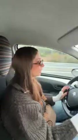 A woman with long brown hair and sunglasses drives a small white car on a highway. She wears a bulky brown sweater and jeans. A passenger sits beside her; only a hand and blue jeans are visible. The car interior is grey and black. The highway stretches ahead, with other vehicles visible in the distance under a cloudy sky.