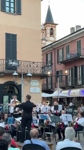 A brass band performs in a town square. A conductor in a black shirt and pants stands facing the band, baton raised. The band members, wearing white shirts and red accents, play various instruments including trumpets, trombones, saxophones, and tubas. They are seated on chairs behind music stands. The audience sits in the foreground. In the background, buildings with balconies and awnings line the square, and a church tower with a spire and bell is visible. The lighting suggests it is late afternoon or early evening.