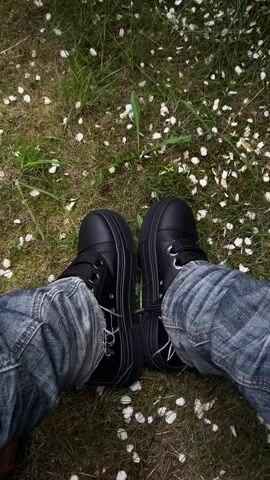 A first-person video shows someone sitting in a blue fabric hammock with their feet in black shoes with thick soles and dark wash jeans resting on the ground, surrounded by green grass and scattered white petals. The camera pans upwards, revealing the blue hammock tied around a tree and a stone fence with a wooden gate in the background.