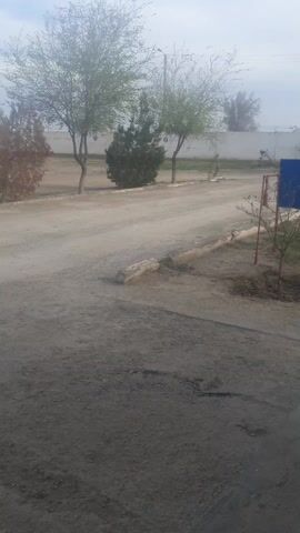 The video shows a dirt road leading away from the camera, with trees lining the sides. A blue sign is visible on the right, near a small tree. A concrete walkway leads to a small white building with a grey roof. The sky is overcast. The camera pans slightly to the right.