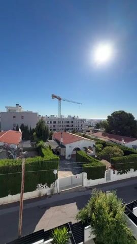 This is a vertical video shot from a high vantage point, looking out over a residential area on a sunny day. The sun is high in a clear blue sky, casting bright light and lens flares. In the foreground, the terracotta tiled roof of a building is visible. Below, a street runs horizontally, lined with white fences and neatly trimmed green hedges. Several houses with red-tiled roofs are visible, interspersed with trees. In the middle ground, a construction crane is prominent against the sky, suggesting ongoing development. Further in the distance, the sea is visible, meeting the horizon under the bright sun. The overall impression is of a calm, sunny day in a coastal residential neighborhood.