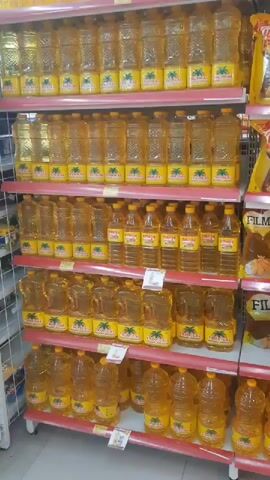 The video shows a grocery store aisle stocked with cooking oil. The shelves are filled with rows of identical bottles and pouches of cooking oil, primarily in large plastic bottles and yellow pouches. The lighting is bright, typical of a retail environment. The camera pans across the shelves, showing multiple brands and sizes of cooking oil. There are no people visible in the video, and the focus is entirely on the products on display. The aisle appears well-organized and fully stocked.