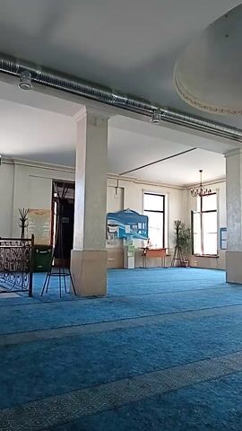 The video shows a room with blue carpeting, likely a mosque. There are pillars, windows, and a chandelier visible. In the background, there is a green mihrab, a pulpit, and bookshelves. The camera is panning and tilting, giving a panoramic view of the space. The lighting suggests it is daytime, and the room appears to be empty.