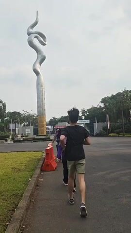 A man in a black t-shirt and khaki shorts walks away from the camera down a sidewalk next to a large silver sculpture. Three people are walking in front of him. They are walking toward a white truck. There are orange barriers on the left side of the sidewalk. The sky is overcast.
