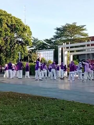 A group of approximately 20 people are performing a synchronized exercise routine outdoors on a paved surface. They are mostly dressed in purple tops and white pants, with some wearing purple headscarves. The individuals are arranged in several rows, facing the same direction, and appear to be engaged in movements such as arm raises and squats. In the background, there are large trees, a modern building with white columns, and a large screen or billboard. The lighting suggests it is daytime, likely morning or late afternoon, with a clear sky. The foreground shows a patch of grass.