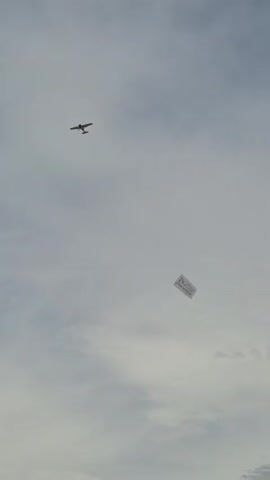 A small airplane flies through a cloudy sky, pulling a banner. The banner is rectangular and appears to have text and possibly a logo on it. In the lower portion of the frame, a beach scene with a few people and structures is visible, though it is largely out of focus. The airplane is in the upper left quadrant of the image, and the banner is to the right and slightly below the airplane. The banner appears to be white with dark text. The sky is overcast with gray clouds.