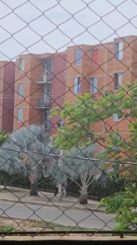 The video shows a view through a wire mesh fence. In the distance, there is a reddish-brown brick apartment building with multiple windows and balconies. The scene includes trees and other greenery, partially obstructing the view of the building. People can be seen walking on the sidewalk near the apartment. In the background, there is a mountain range against a cloudy sky. The camera moves slightly, panning across the scene.