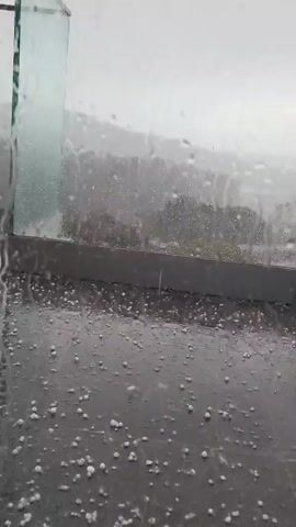 The video shows a view through a glass pane during a hailstorm. The pane is covered with raindrops and streaks. Outside, hailstones cover a dark grey or black surface, possibly a balcony or ground. The hailstones appear round and white. The background is a blur of grey sky and indistinct landscape features, likely trees or hills obscured by the weather. The hailstorm seems intense, with visible movement of hail and rain.