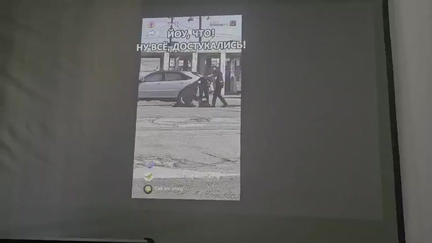The video shows a projected image on a screen. The projected image is a vertical video feed, possibly from a social media platform. The top of the video displays text in Russian. In the background of the video, a silver sedan is parked on a street. A person, appearing to be a police officer in uniform, is interacting with a large dark-colored dog. The officer is crouched down, and the dog is in front of them. Below this scene, there are colored circles with icons and text in English. The second frame shows a close-up of a police car's wheel and side mirror. The subsequent frames show the police officer running towards the camera and then striking a pose with their arms outstretched. The text at the top of the projected image appears to change between frames, also in Russian. The environment appears to be an outdoor urban setting, with asphalt pavement and some buildings visible in the distance. The projection itself is slightly blurry and washes out in some areas.