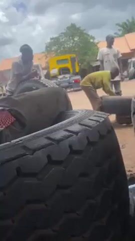 The video depicts four men in an outdoor setting with a truck and several large tires. A man sits on a pile of tires on the left side of the frame. Another man stands beside the tires, while a third person bends over a tire to the right. In the background, a man is holding something in his hands. A yellow truck cab is visible in the middle of the scene, and a building with a red tile roof is in the far background. The ground appears to be sandy, and the sky is cloudy.
