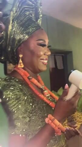 The video shows a woman dressed in traditional Nigerian attire. She is wearing a green, shiny dress, a matching head wrap, and orange coral necklaces and bracelets. She is holding a white object, possibly a small speaker or microphone, and smiling while she speaks. The background is a green wall, with a glimpse of a framed picture and a door. Another person, partially visible, stands to her left. The overall setting suggests a celebratory event, possibly a wedding or traditional ceremony.