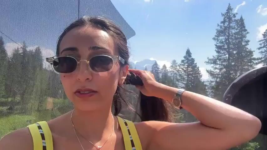 A woman wearing sunglasses and a yellow top is riding in a cable car. She is holding her hair in her right hand. The cable car has a mesh covering over the window, and through the window, a forest and mountains can be seen. The woman appears to be speaking to the camera.