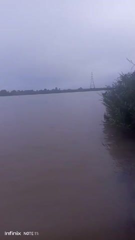 The video shows a body of water, possibly a river or lake, under a grey and overcast sky. The water appears murky and brown. Along the edge of the water, there is some vegetation. In the distance, the horizon line is visible with a faint outline of trees and structures. There is a tower-like structure in the distance. The video seems to be taken from a low vantage point, close to the water's edge. The scene has a quiet, slightly ominous feel due to the grey, muted colors and the wide expanse of water.  The words "Infinix NOTE 11i" are visible in the bottom left corner.